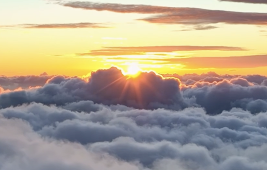 The sun rises behind a cloud, casting warm orange and yellow light over a dense blanket of fluffy clouds in the sky—an inspiring scene reminiscent of NEOM’s visionary landscapes.